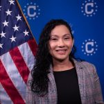 Person in a plaid blazer standing before a blue backdrop with U.S. government emblems and beside an American flag with visible stars and red‑white stripes