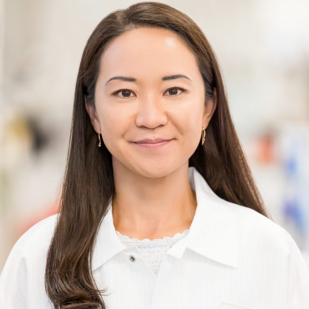 a person with long, dark hair wearing a white lab coat. The lab coat has a name tag on the left side that reads "NMS" with a blue triangular logo above the text. The background is blurred, making it difficult to discern specific details, but it appears to be an indoor setting.