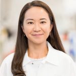a person with long, dark hair wearing a white lab coat. The lab coat has a name tag on the left side that reads "NMS" with a blue triangular logo above the text. The background is blurred, making it difficult to discern specific details, but it appears to be an indoor setting.