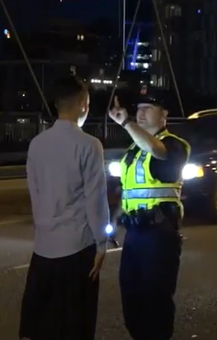 Officer in reflective yellow vest conducts nighttime sobriety test on man along city street, illuminated by car headlights
