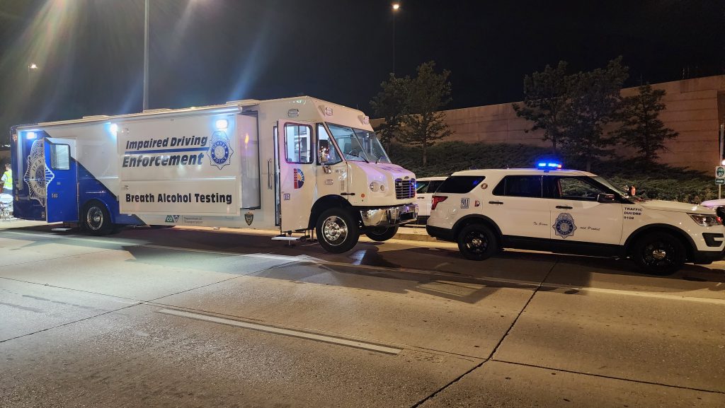 White truck marked 'Impaired Driving Enforcement' and 'Breath Alcohol Testing' parked at night with police SUV behind, lights flashing