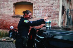 An officer places decontamination bottles on a patrol vehicle’s hood beside a brick building, with a person seated on the ground near the wall