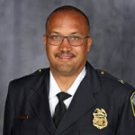 Uniformed police official wearing dark dress coat with gold badge, black tie, white shirt, shoulder epaulettes, and department shoulder patch, posed against mottled gray studio background.