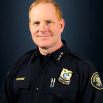 Person wearing a dark navy Portland Police dress uniform with gold badge reading “Deputy Chief,” collar stars, nameplate, pens in pocket, and shoulder patch, posed against a dark blue backdrop.