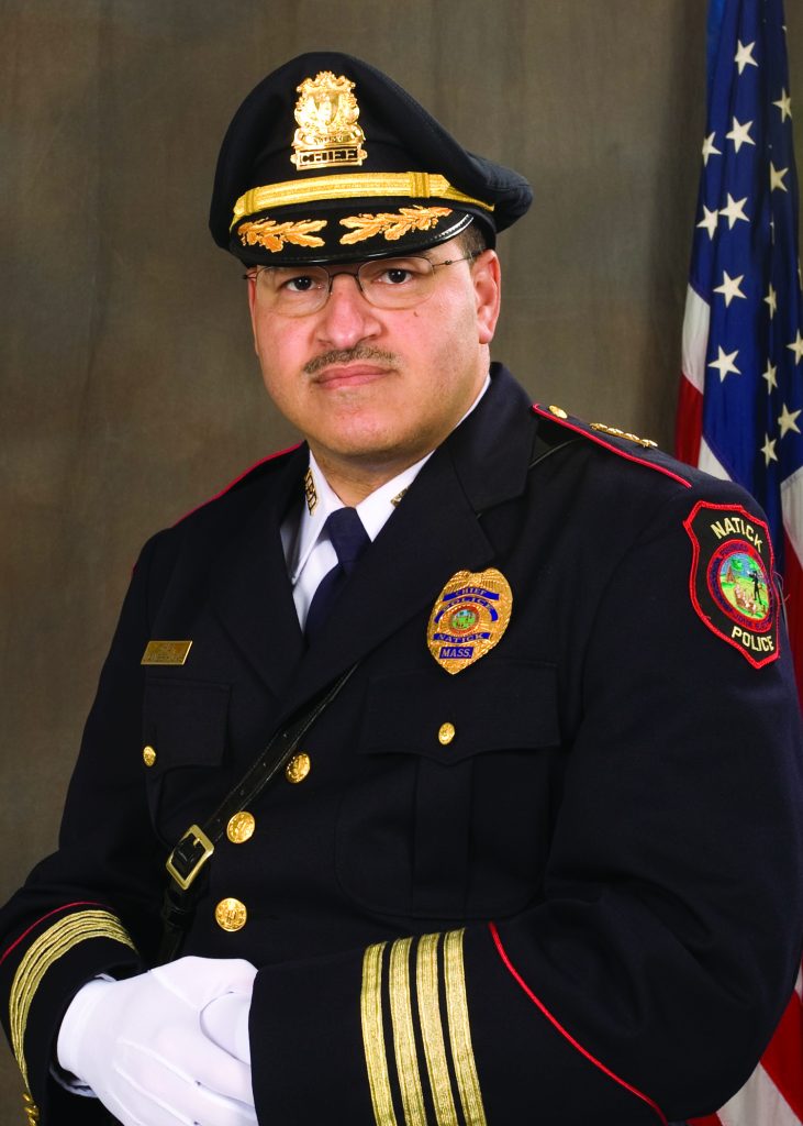 Police chief in formal dark uniform with gold buttons, badge, shoulder patch reading Natick Police, white gloves, and peaked cap, standing before U.S. flag.