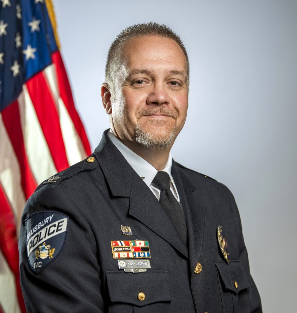 Police chief in dark formal uniform with gold buttons, badge, service ribbons, and Salisbury Police patch, posed in front of U.S. flag and gray backdrop