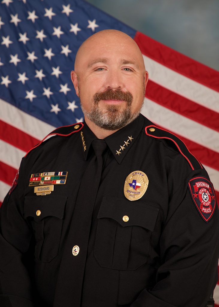 Uniformed police chief wears a dark dress uniform with gold badge, ribbons, collar stars, shoulder patch, and nameplate, posed before a U.S. flag backdrop