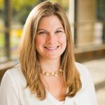 Individual with shoulder-length blonde hair in white blouse and pearl necklace stands indoors; blurred windows and greenery visible in background