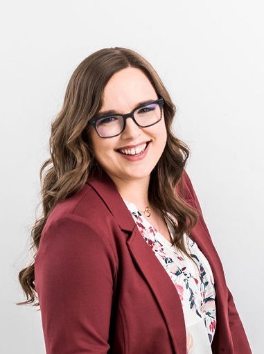 Individual wearing glasses, maroon blazer, and patterned blouse stands at an angle against a plain gray studio background, with long wavy hair and soft lighting