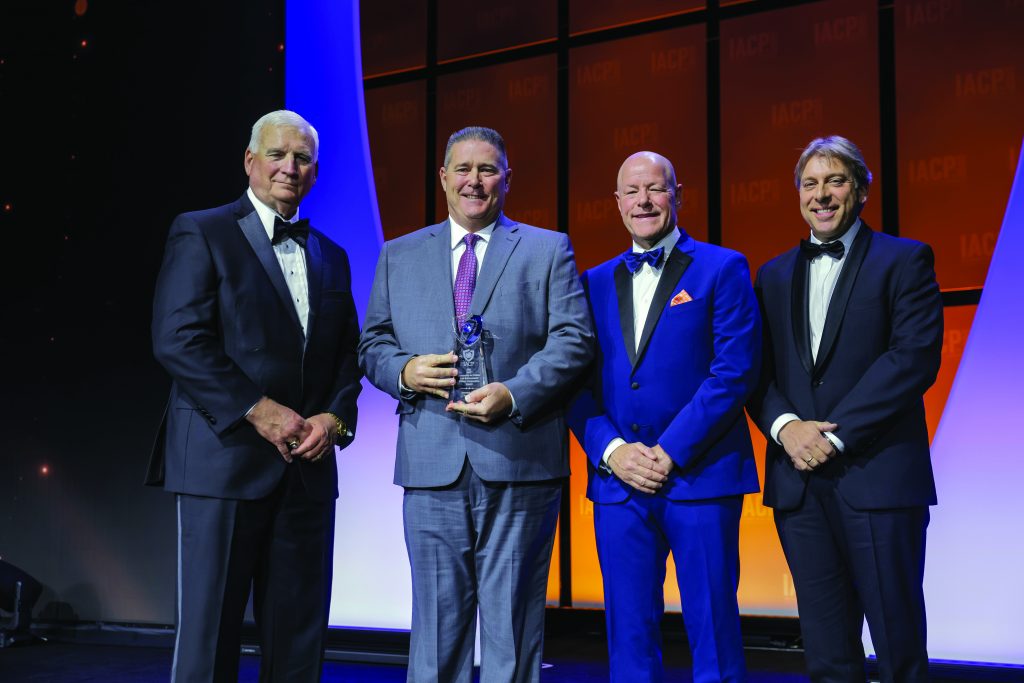 Four individuals in formal attire stand on stage with orange IACP backdrop; one in gray suit holds a clear glass award, others wear black tuxedos and a blue suit.