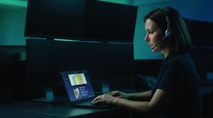 Communications operator wearing a headset works at a laptop displaying maps and incident data, surrounded by multiple dark monitors in a low‑light command center environment