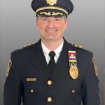 Police chief in dark dress uniform with gold buttons, badge, shoulder patches, rank stars on collar, service ribbons, and peaked cap against gray backdrop.