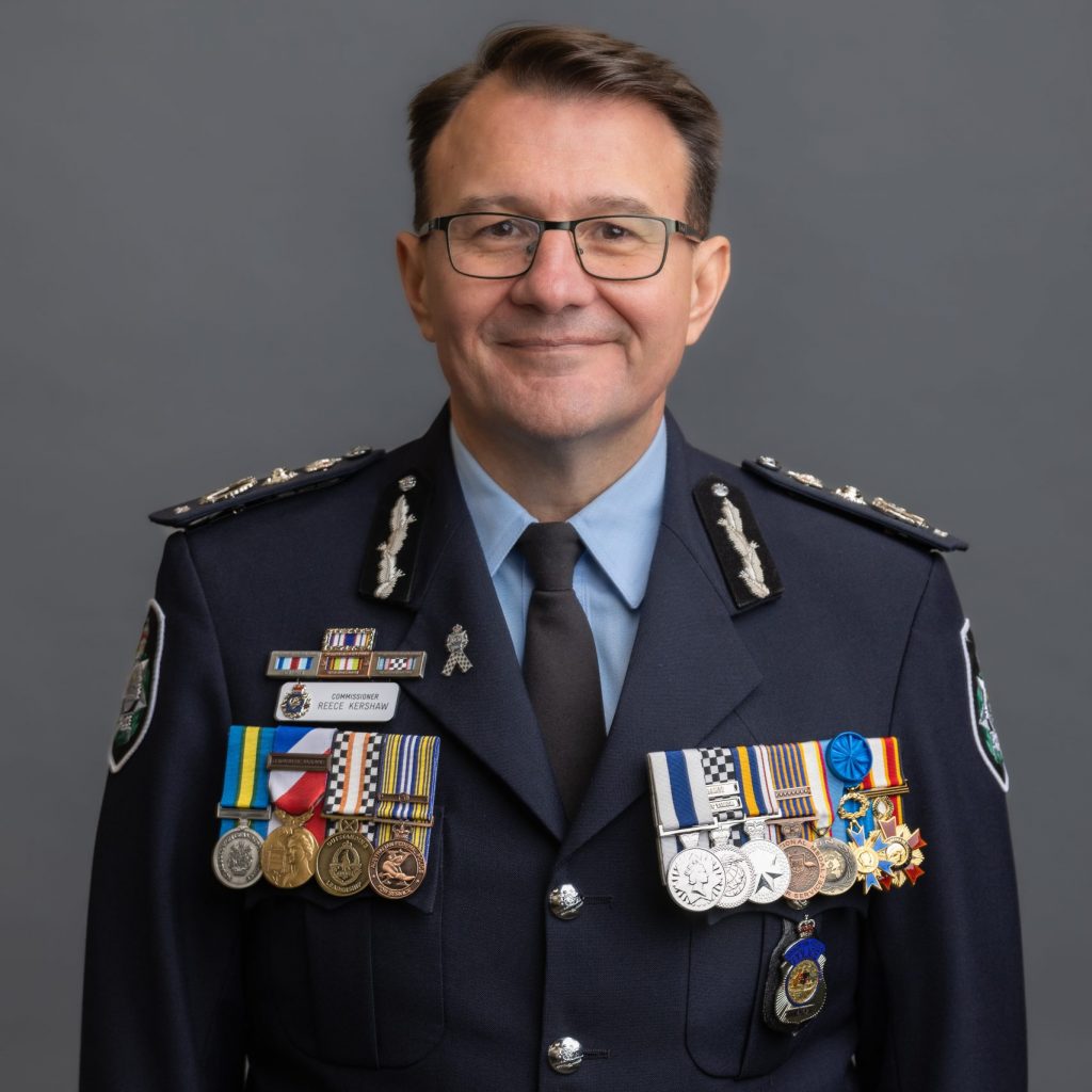 officer in decorated formal uniform with medals, badges, and name tag “M. Huxley,” standing before a plain gray backdrop