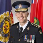 Ontario Provincial Police officer in uniform with peaked cap, medals, and shoulder insignia stands between OPP flag and Ontario flag