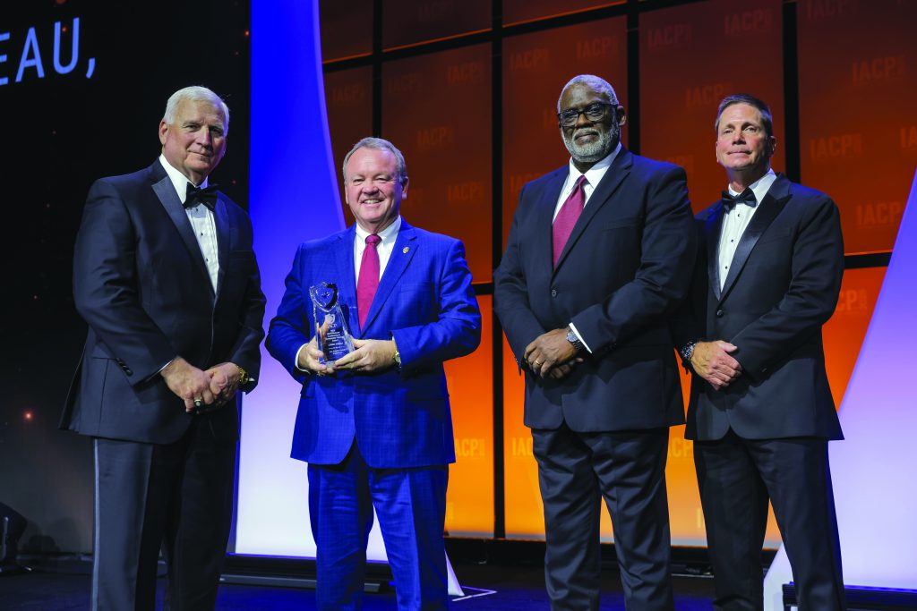 Four individuals in formal attire stand on stage with orange IACP backdrop; one in a blue suit holds a clear glass award, others wear black suits with ties.