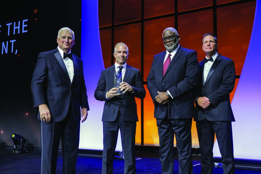 Four individuals in formal attire stand on stage with orange IACP backdrop; one in dark suit holds a clear glass award with blue accent, others wear tuxedos.