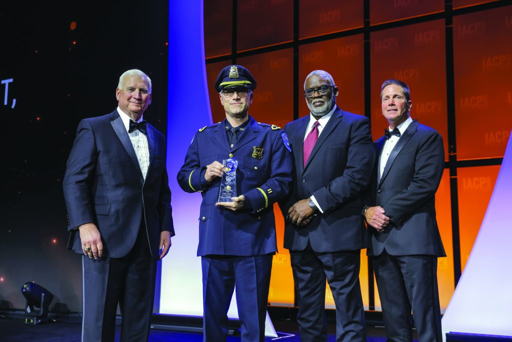 Four individuals stand on stage with orange IACP backdrop; center person in dark police dress uniform and peaked cap holds a clear glass award with blue accent.
