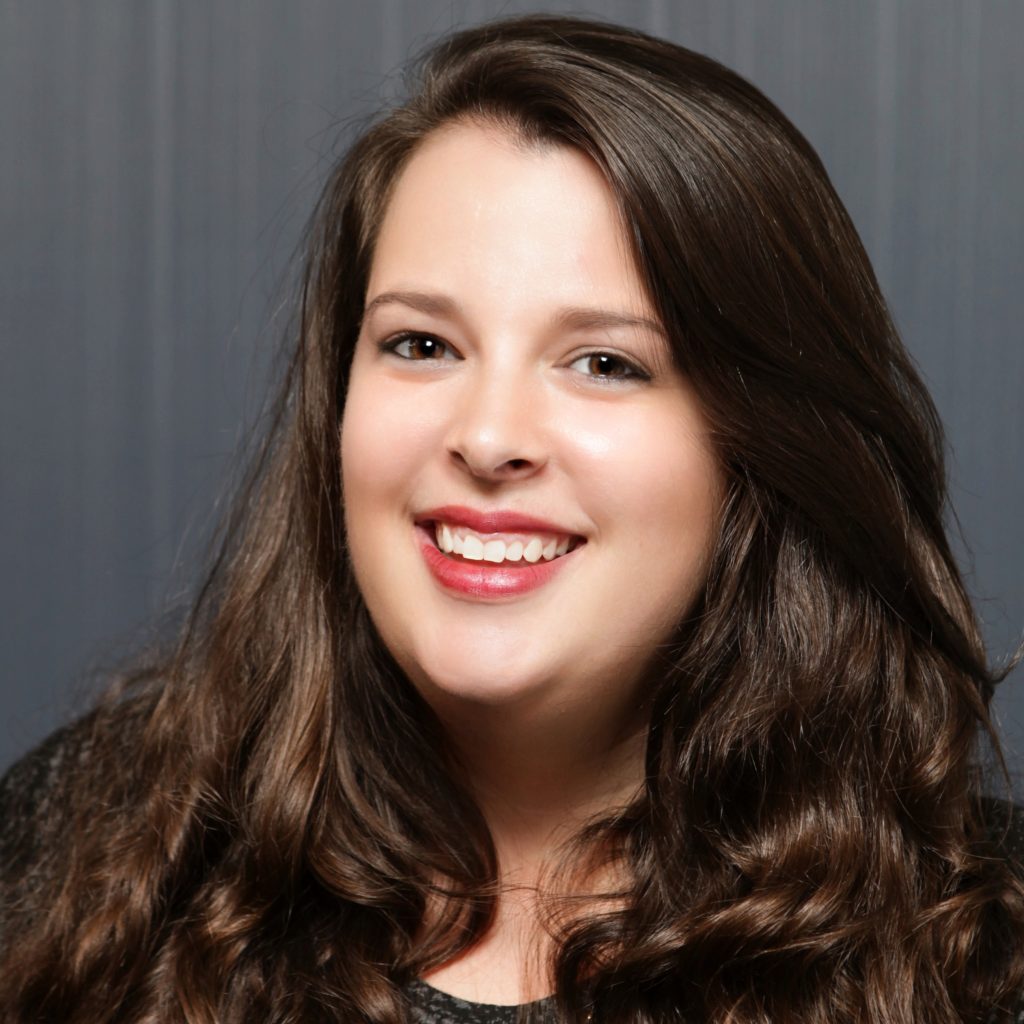 Person with long wavy brown hair wearing a dark textured top stands before a gray vertical-paneled backdrop in a studio-style portrait setting.