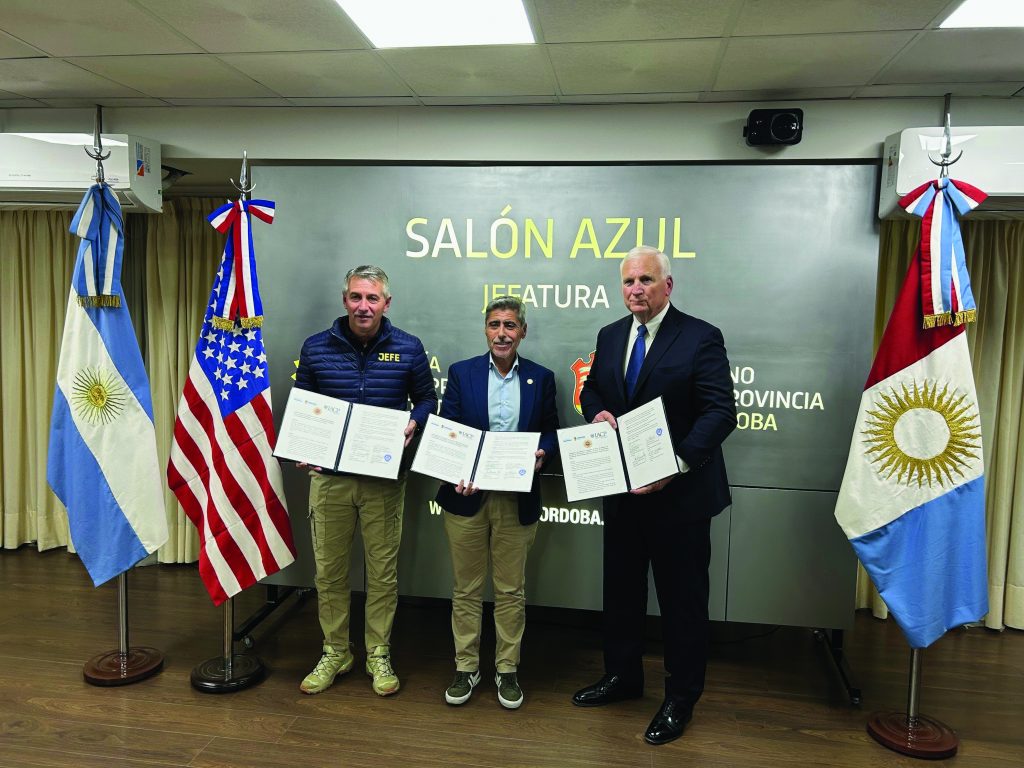 Three individuals hold documents in front of 'SALÓN AZUL JEFATURA' backdrop, flanked by Argentine and US flags, indicating formal signing event.