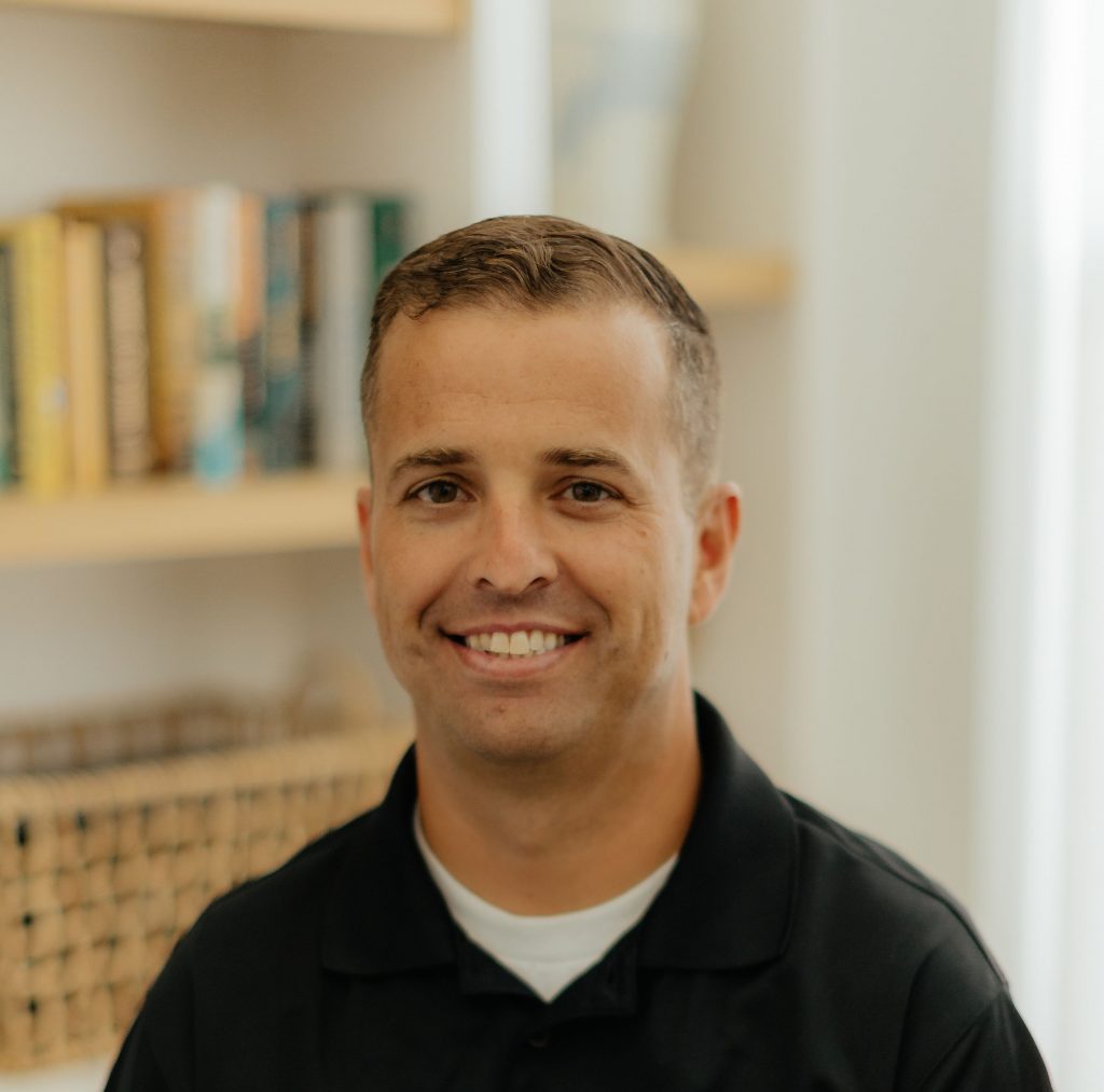 An individual wearing a black collared shirt is seated indoors with a light-colored wall, wooden shelves holding books, and a woven basket visible in the background