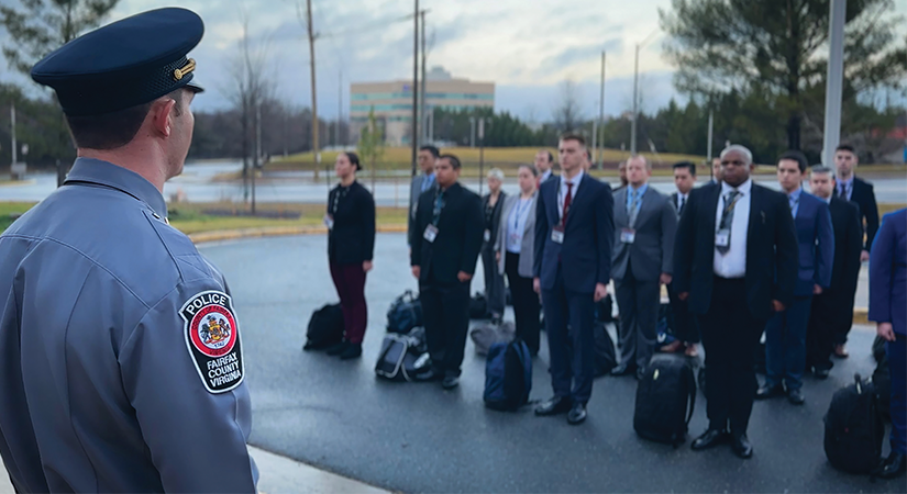 Fairfax County Police officer in uniform faces line of formally dressed individuals outdoors, each wearing ID badges and carrying backpacks