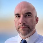 Person in white dress shirt and blue tie stands before blurred outdoor backdrop with blue sky and beige terrain; face intentionally obscured