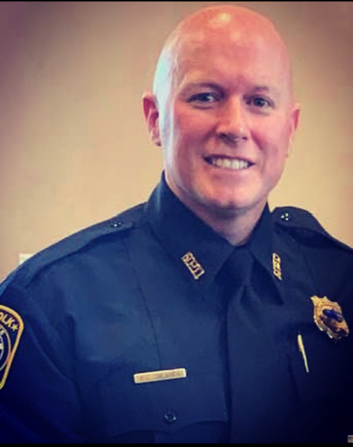 Studio portrait of a uniformed police officer wearing a dark dress uniform with shoulder patches, badge, nameplate, collar rank pins, and tie, posed against a neutral indoor background