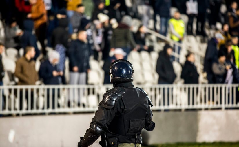 Riot‑gear officer in helmet and body armor stands facing a crowd behind white barriers in stadium seating area, with high‑visibility staff and rows of empty seats