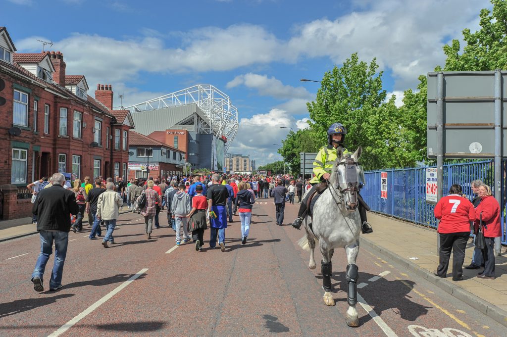Mounted officer in high‑visibility jacket rides a gray horse along a crowded street of fans heading toward a stadium, with brick homes, fencing, signage, and blue sky