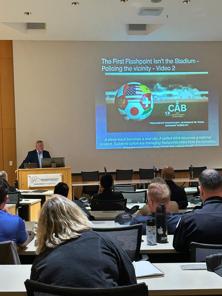 Instructor at podium presents slide titled ‘The First Flashpoint Isn’t the Stadium’ to classroom audience seated at desks with laptops, bottles, and notes