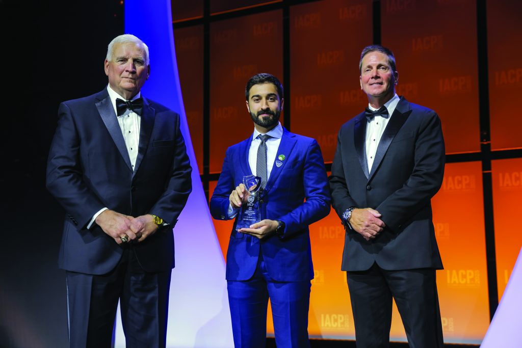 Three individuals stand on stage with orange IACP backdrop; center person in bright blue suit holds a clear glass award with blue accent, others wear black tuxedos.