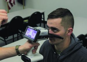 Handheld DAX Evidence Recorder captures close-up eye imagery during a sobriety assessment, with live display screen, alignment arm, examiner’s hand, classroom tables, chairs, and equipment visible