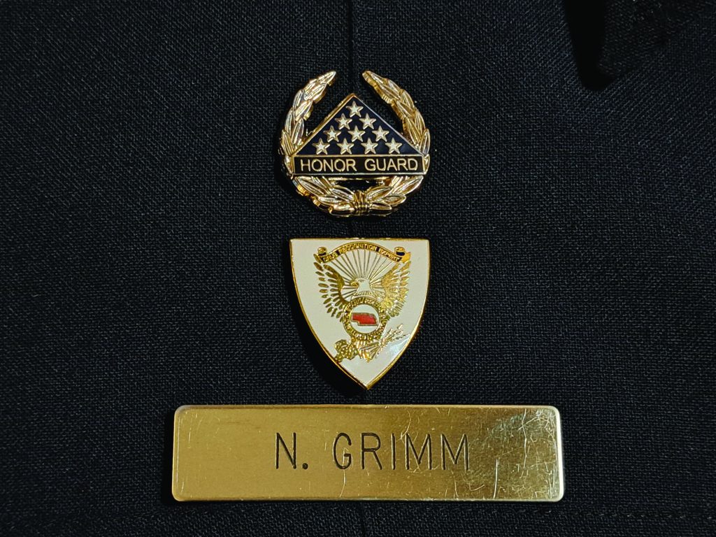 Close-up of law enforcement uniform with 'Honor Guard' badge, eagle emblem shield, and nameplate reading 'N. Grimm'; dark fabric background
