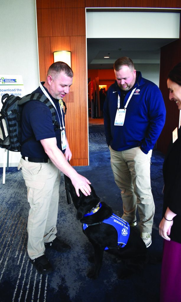 Two individuals wearing conference badges and casual attire stand indoors; one pets a black service dog in a blue vest marked “Service Dog” near a wooden wall panel.