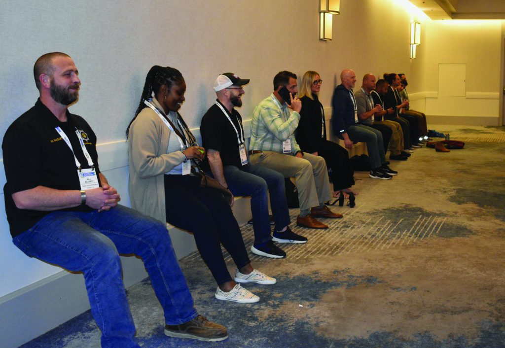 The image shows eleven individuals participating in a wall sit exercise in a hallway with beige walls and carpeted flooring. They are lined up side by side, leaning against the wall with their backs straight and knees bent at a 90-degree angle, as if sitting on invisible chairs. Each person is wearing a conference badge around their neck, indicating they are attending an event. The participants are dressed in various attire, including casual clothing such as jeans and sneakers, as well as business casual outfits like slacks and blouses. Two wall-mounted lights illuminate the area above them. On the floor to the right side of the image, there is a red bag or equipment case placed near the participants.