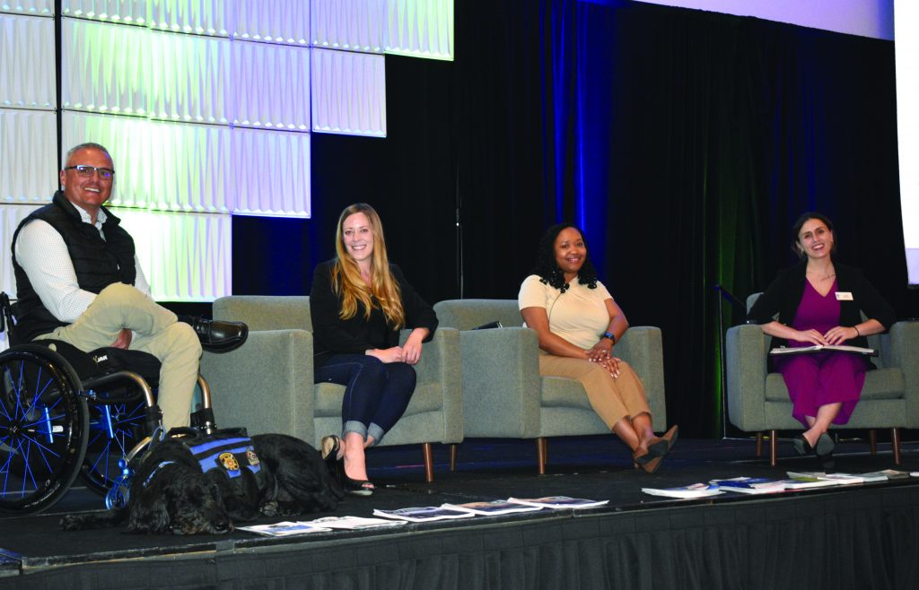 a panel of four individuals seated on a stage, each in separate armchairs. From left to right: the first individual is a person in a wheelchair with a service dog lying down in front of the wheelchair. The second individual is sitting with legs crossed and hands resting on their lap. The third individual is also seated with legs crossed and hands resting on their lap. The fourth individual is holding some papers or notes, possibly moderating or taking notes for the discussion. Behind them, there are large illuminated panels forming part of the backdrop, and dark curtains framing the stage area. There are several documents or booklets placed on the edge of the stage floor in front of them.