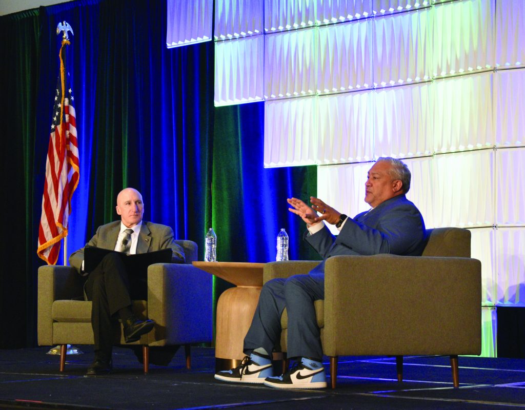 two individuals seated on stage in a formal setting, likely during a panel discussion or interview. Both individuals are sitting in gray armchairs with a small wooden table between them, which holds two water bottles. The individual on the left is dressed in a suit and tie, while the individual on the right is wearing a blue suit and white sneakers with black accents. Behind them is an American flag positioned to the left side of the stage. The backdrop consists of large panels illuminated with blue and green lighting, creating a professional atmosphere suitable for conferences or formal events.