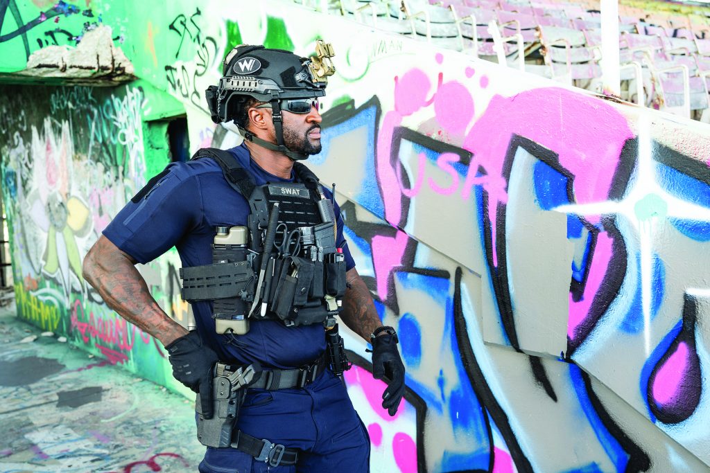 A person in tactical gear and helmet stands beside a graffiti‑covered concrete wall in an abandoned structure, wearing a vest with equipment and a holstered tool
