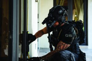 A tactical‑gear‑equipped individual kneels at a doorway, using gloved hands to work with tools on the door frame while wearing a helmet, vest, and mounted equipment