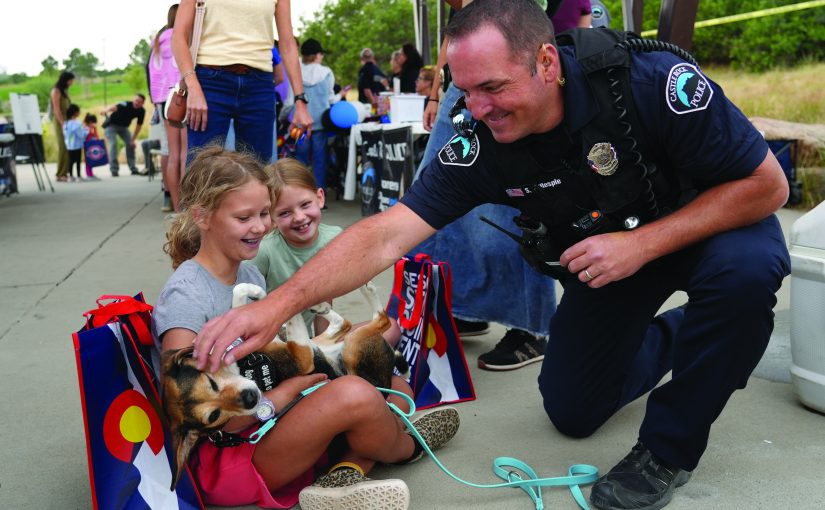 Uniformed officer kneels beside two children holding a dog at an outdoor community event, with tactical vest, badge, radio, booths and crowd behind.