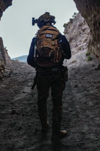 Soldier in camo gear and helmet with mounted camera stands at rocky cave entrance, facing out; carries large tan backpack with visible patches.