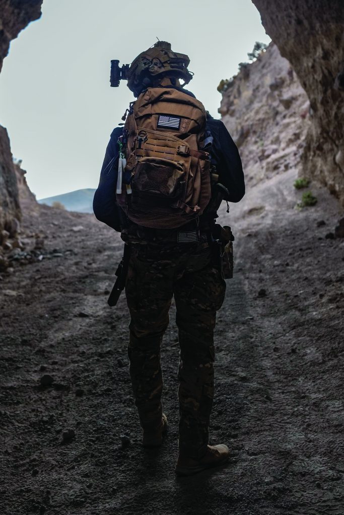 Soldier in camo gear and helmet with mounted camera stands at rocky cave entrance, facing out; carries large tan backpack with visible patches.