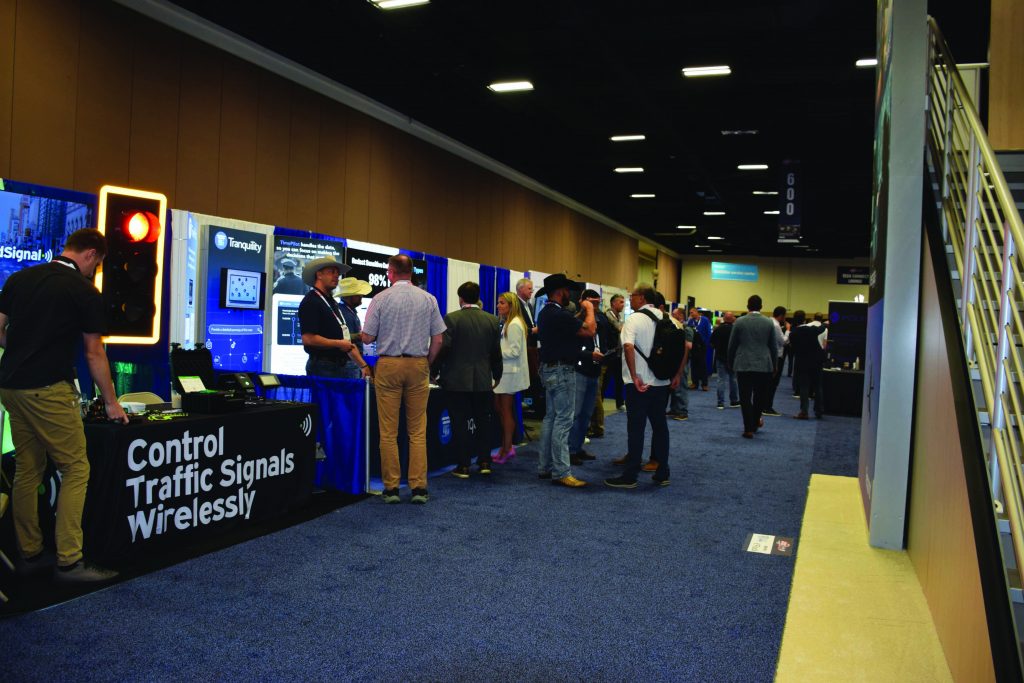 Conference expo hall with attendees in business attire browsing booths, signage reading ‘Control Traffic Signals Wirelessly,’ monitors, lighting panels, and vendor displays lining the aisle