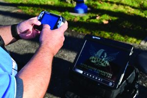 Person holding handheld device with blue screen near mounted rugged tablet displaying Patrol PC logo outdoors on pavement with grass in background