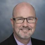 Person wearing a dark suit jacket over a light pink collared shirt, posed against a smooth gray studio background with soft lighting