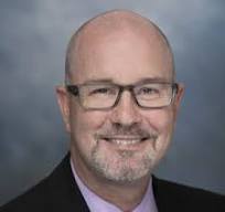 Person wearing a dark suit jacket over a light pink collared shirt, posed against a smooth gray studio background with soft lighting