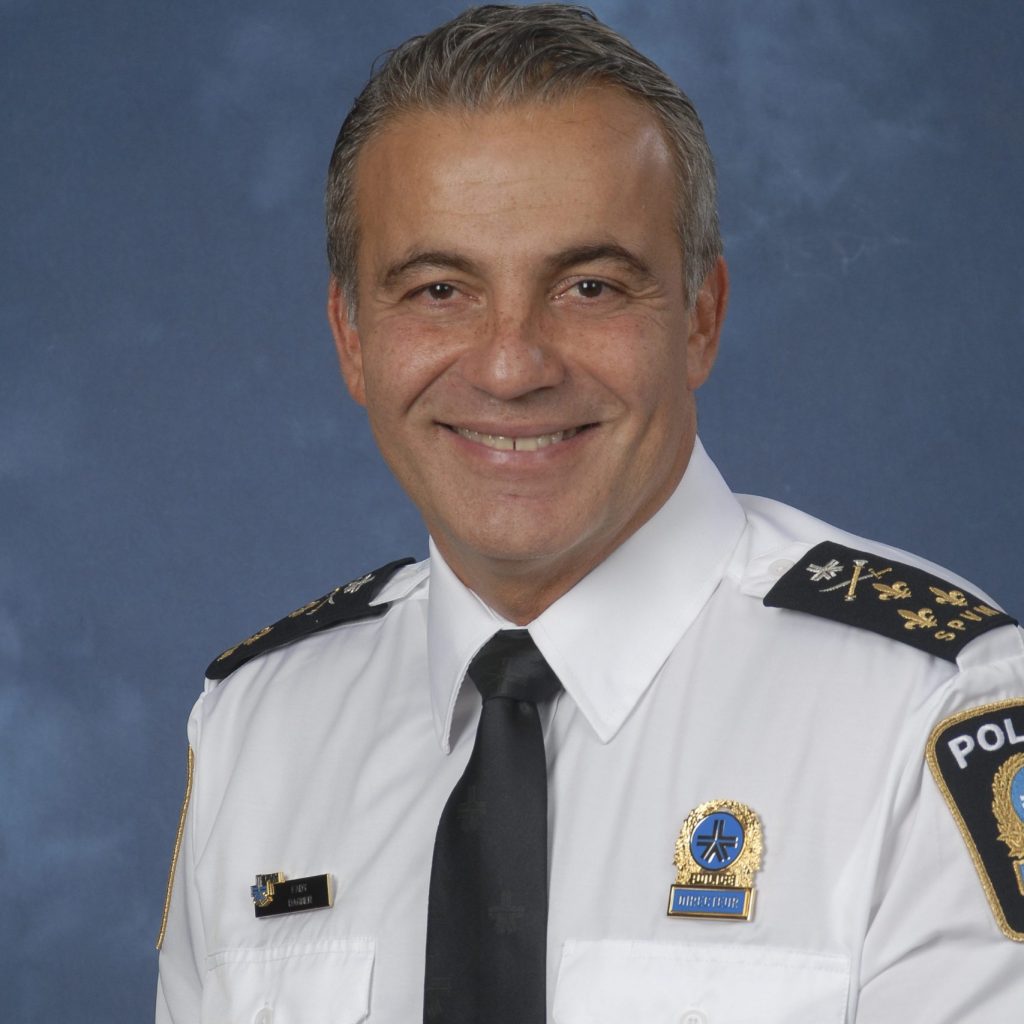 Officer in white uniform with black epaulets bearing gold insignia, black tie, and visible police badges on chest and shoulders—formal police attire