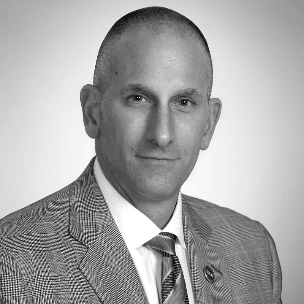 Grayscale image of person in checkered suit, white shirt, striped tie, standing against plain light background; focus on formal attire and composition
