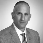 Grayscale image of person in checkered suit, white shirt, striped tie, standing against plain light background; focus on formal attire and composition