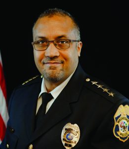 Uniformed officer in dark jacket with gold shoulder stars and badge. Partial American flag visible in background against neutral backdrop